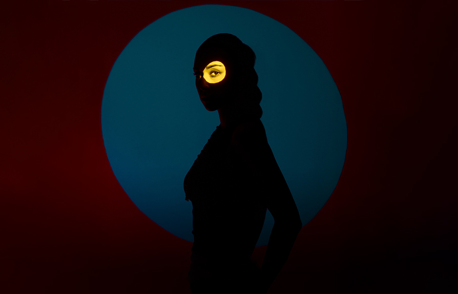 Model posing in studio for abstract photo on a red background with blue spotlight effect and yellow circle accent on her eye made with Godox BFP Flash Projection Attachment using color gels